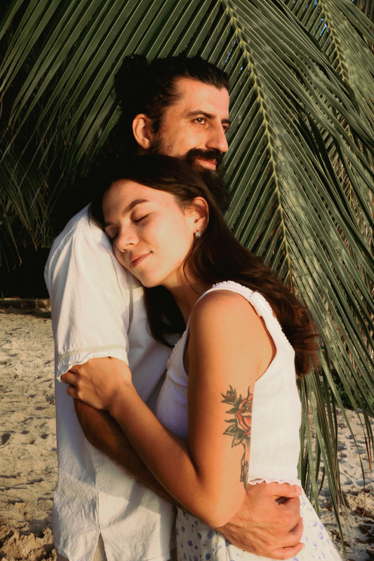 A Couple Hugging Each Other While Standing On The Shore Near Palm Leaves Photos By Canva 